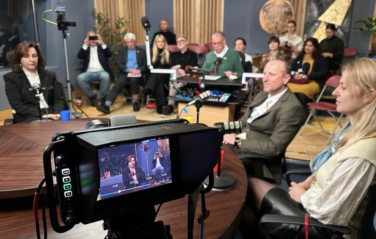A camera records a roundtable discussion in the United Action TV studio, with guests around a wooden table and an audience behind